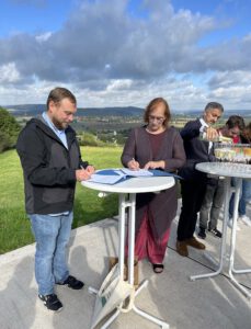 Geschäftsführer Jan Waitzmann und Seminarleiterin Anke Lüth stehen gemeinsam an einem Stehtisch, Beide blicken auf den vor ihnen auf dem Tisch liegenden Kooperationsvertrag und unterschreiben.