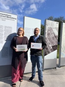 Seminarleiterin Anke Lüth und Geschäftsführer Jan Waitzmann vor Ausstellungstafeln am Dokumentations- und Lernort Bückeberg. Beide blicke in die Kamera. Sie halten jeweils ein Schild in der Hand mit dem Logo ihrer Organisationen.
